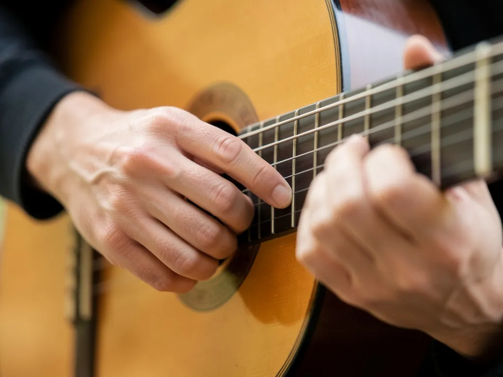Mão esquerda de violonista fazendo pestana com dedo indicador inclinado e polegar como apoio, iluminação suave e clima de concentração